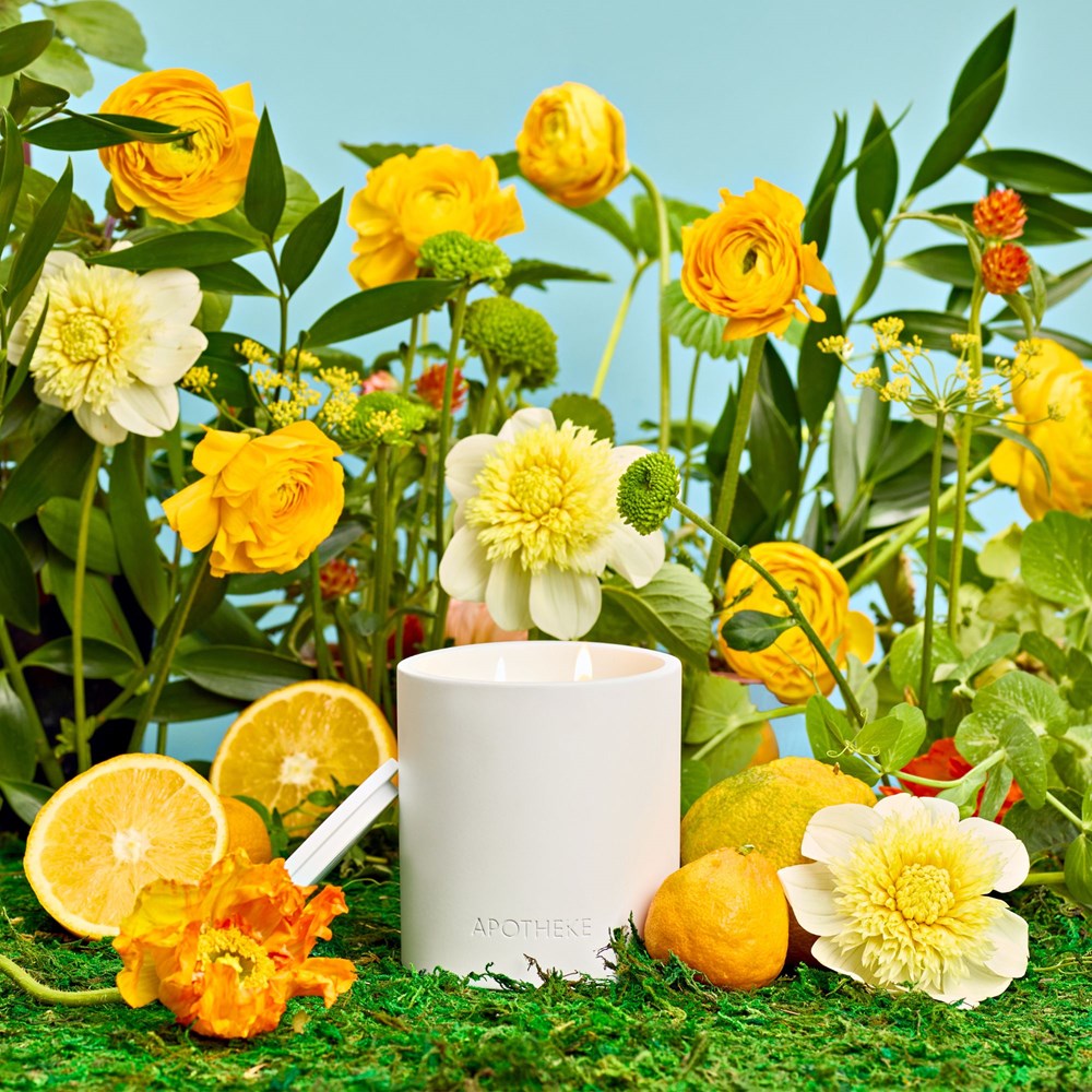 White candle with floral design surrounded by yellow flowers and fruits on a green surface.