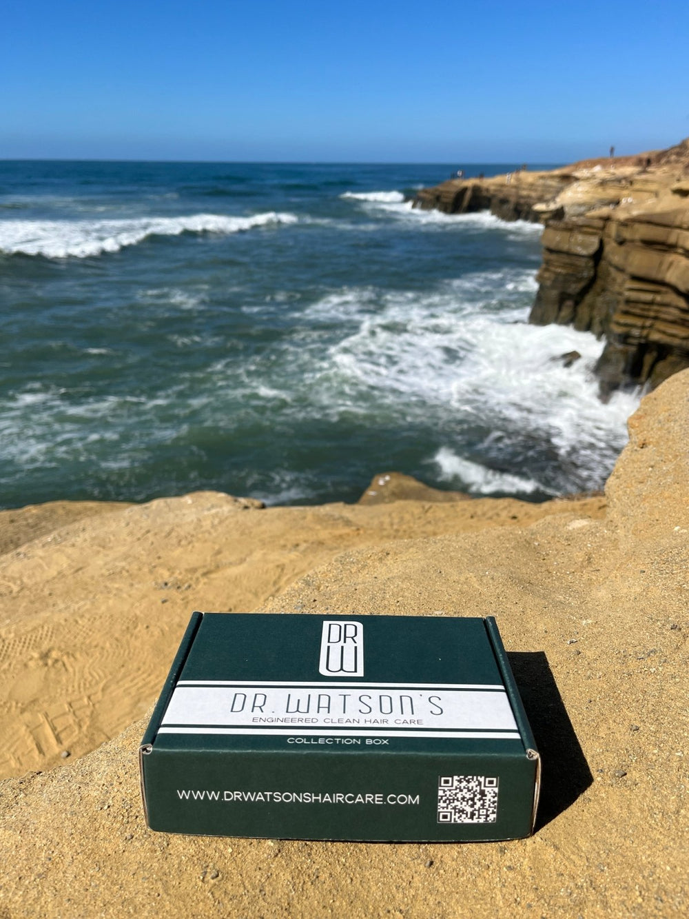 Dr. Watson's hair care collection box on a beach with ocean waves in the background