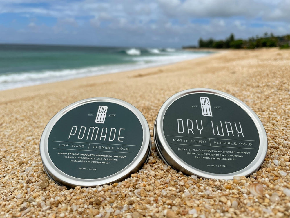 Two containers of hair care products, Pomade and Dry Wax, on a sandy beach with ocean in the background.